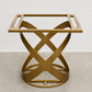 A modern square side table with a gold-finished metal frame and an open, sculptural base made of interlocking curved bands, shown on a light gray concrete floor against a white wall.