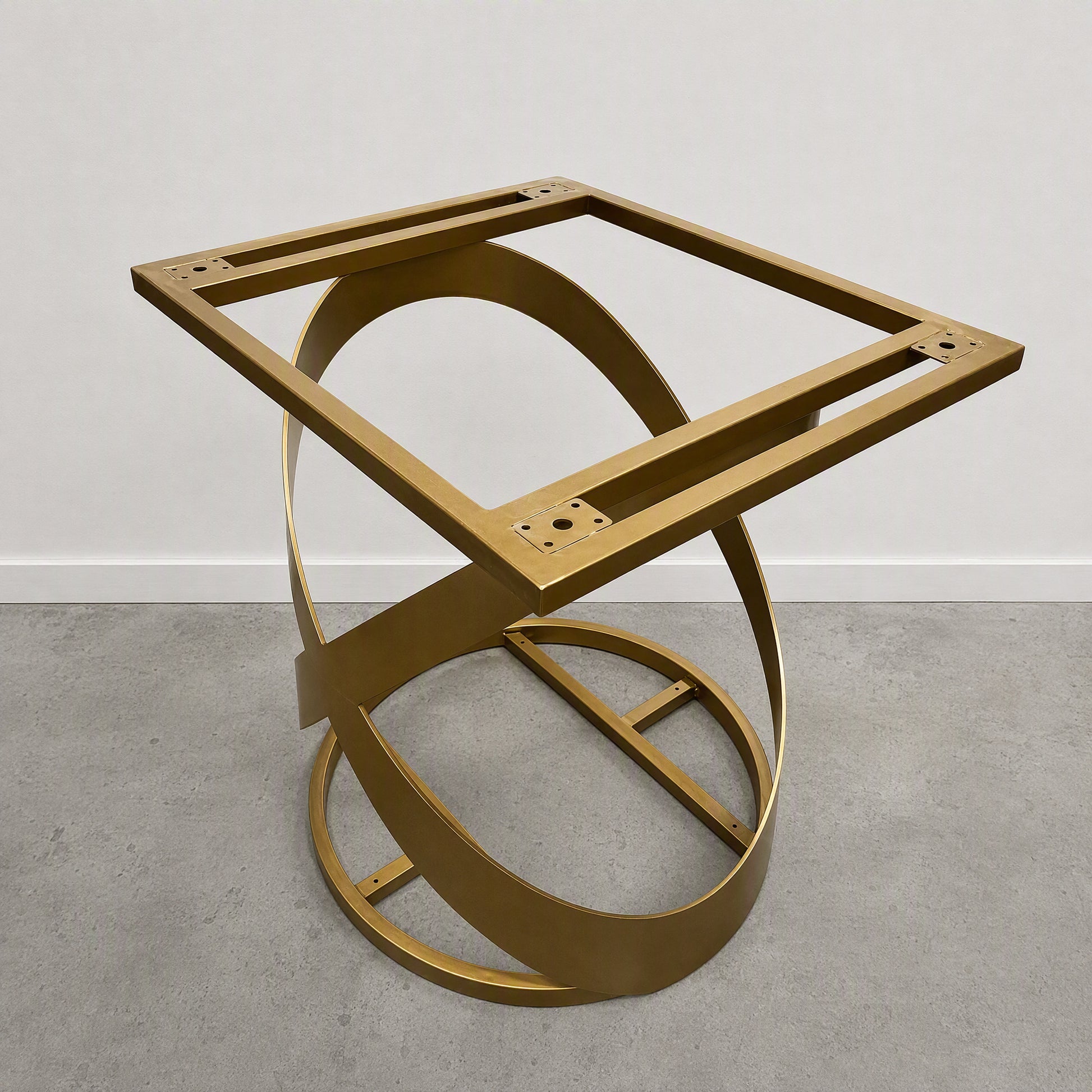 A modern square metal table base with a gold-finished frame and interlocking circular rings, photographed against a plain white wall and gray concrete floor.