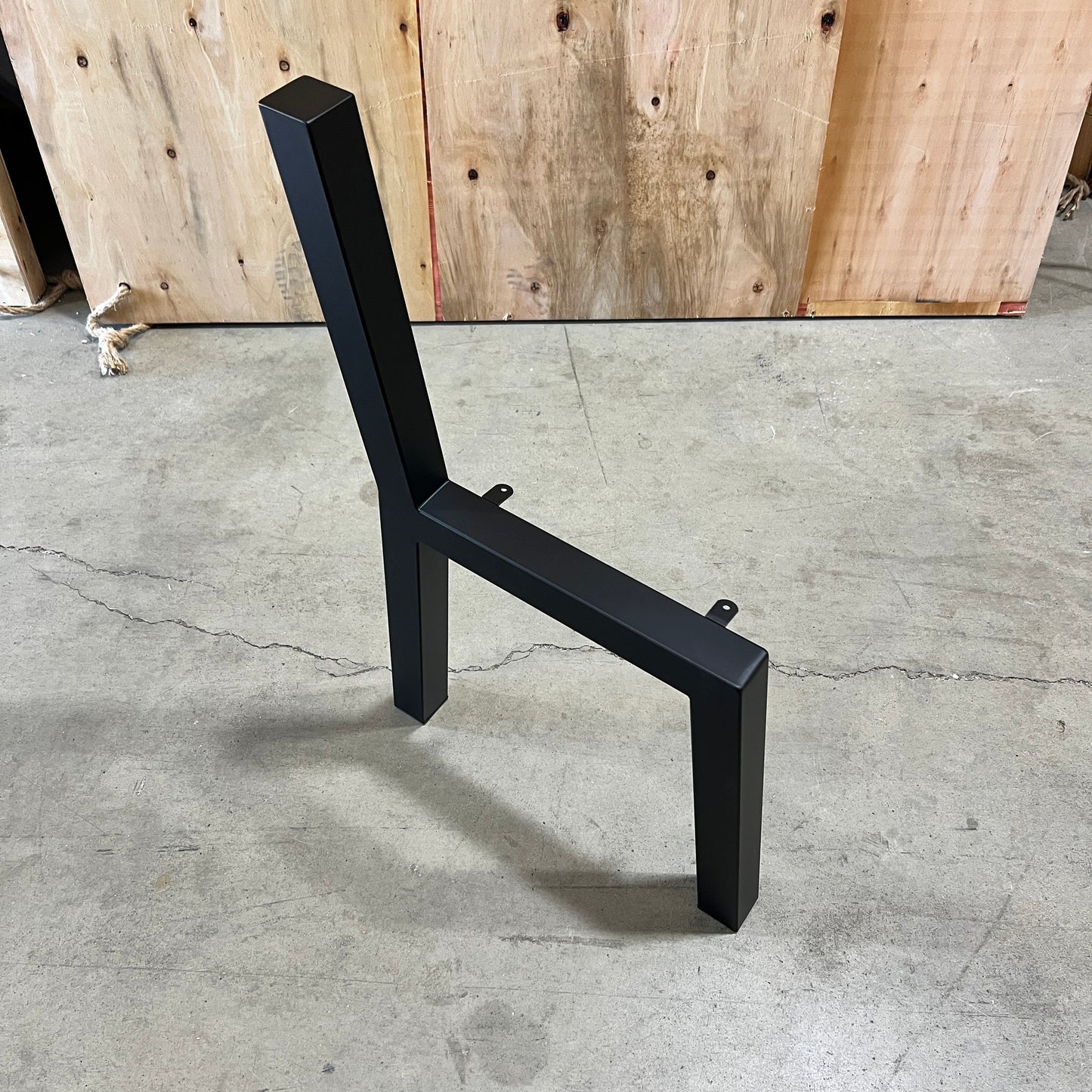A black metal chair frame or table base with a square backrest and horizontal support beam, photographed on a concrete floor in front of a wooden wall.