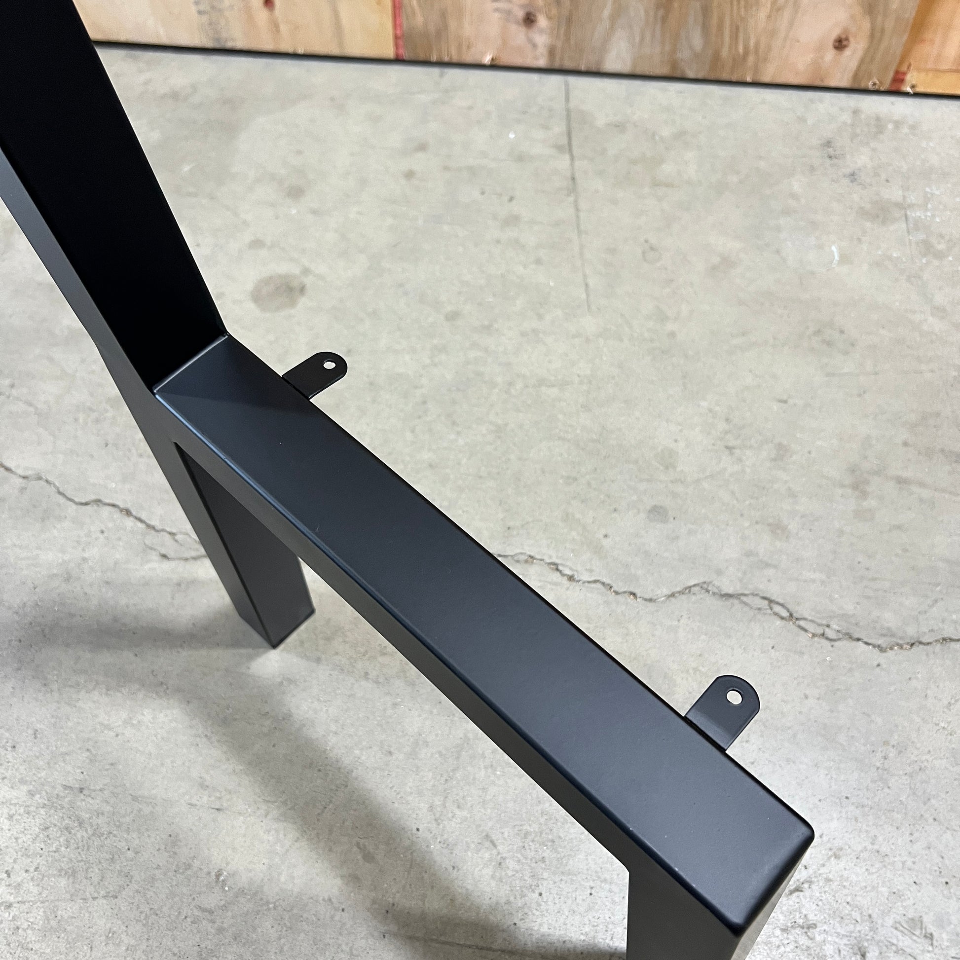 A close-up of a black metal table base or stand leg assembly on a concrete floor, with a wood panel visible in the background.
